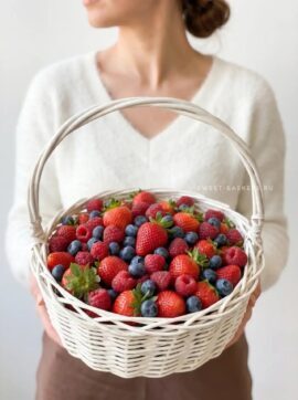 Person in a white sweater holds a white wicker basket overflowing with ripe strawberries, raspberries, and blueberries.