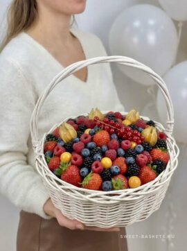 Person in a white sweater holding a white wicker basket overflowing with mixed berries (strawberries, blueberries, raspberries, blackberries, physalis). In the background, pale balloons are visible.