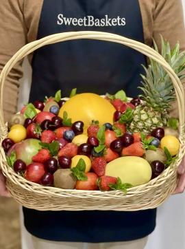 Person in a SweetBaskets apron holds a large wicker fruit basket overflowing with pineapple, mangoes, strawberries, cherries, blueberries and kiwi.