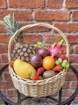 Fruit basket with pineapple, mango, pear, dragon fruit, grapes, strawberries, kiwifruit and oranges in front of a brick wall.