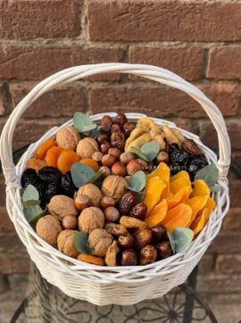 White wicker basket filled with mixed nuts (walnuts, almonds, hazelnuts) and orange dried apricots, with eucalyptus leaves, against a brick wall]