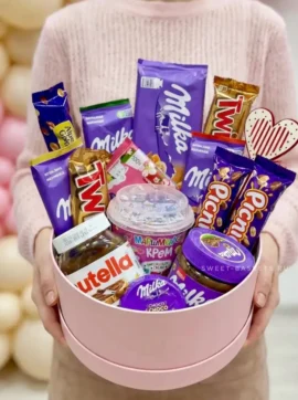 A pink round gift box filled with assorted chocolates and treats, including Nutella jar, Milka bars, Twix, and a jar of cream, held by a person in a sweater.